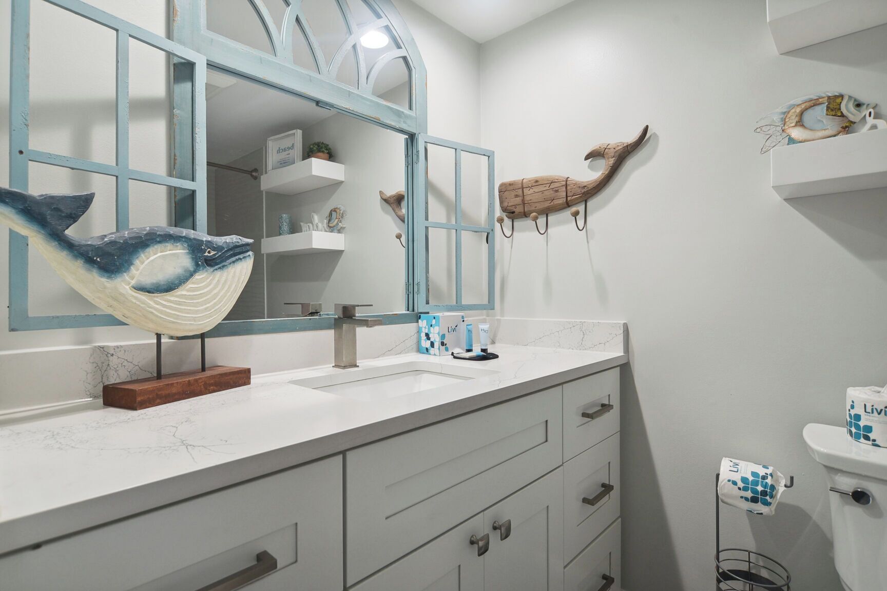 Un baño con un espejo grande, un lavamanos blanco y un inodoro. Hay elementos decorativos como una escultura de ballena y una escultura de pez en la pared.