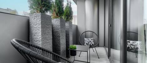 Un balcón con una silla de mimbre, una mesita con una planta y un cerramiento de vidrio que da a una cama.