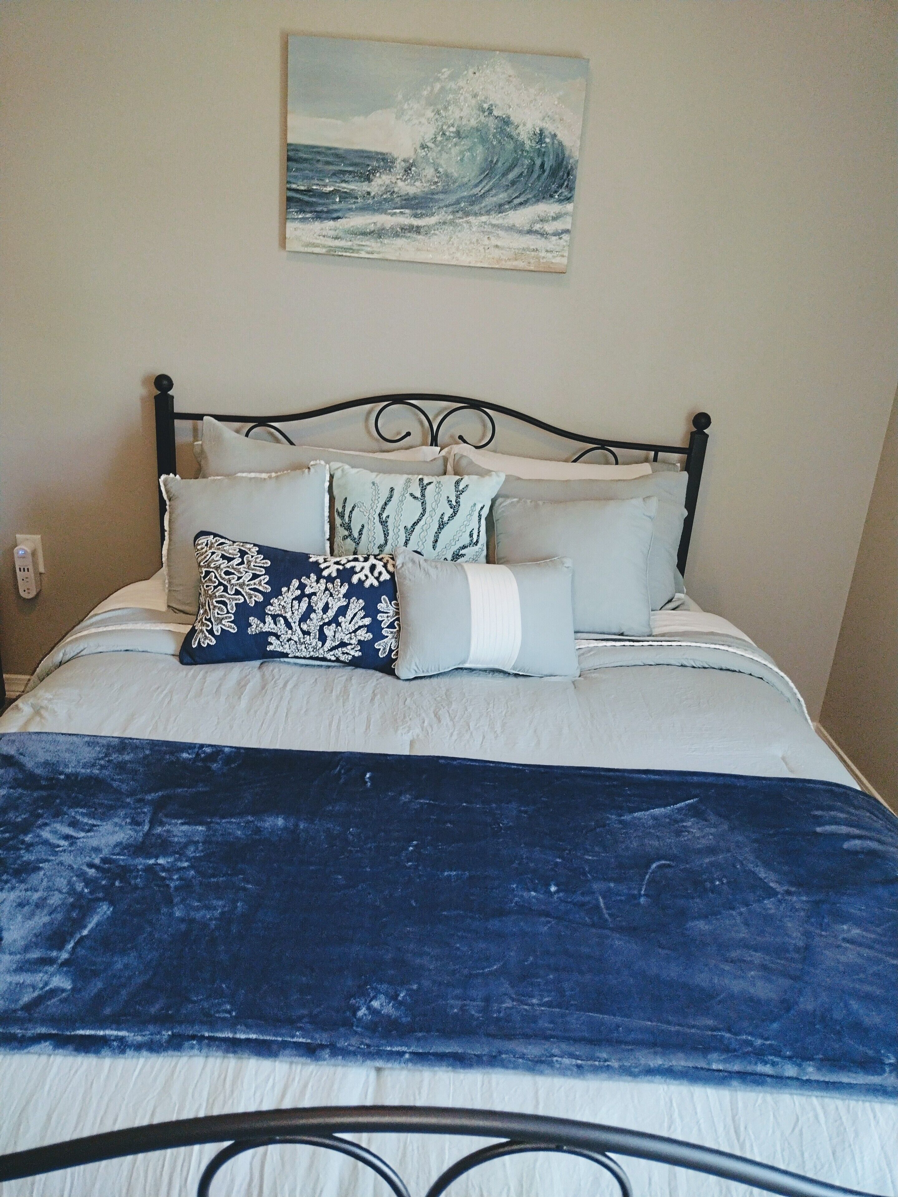 A bed with a dark blue blanket, multiple pillows, and a painting of a wave on the wall.