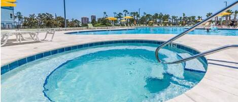 Una piscina con agua azul cristalina, rodeada de sillas de descanso y sombrillas, con palmeras y edificios al fondo.