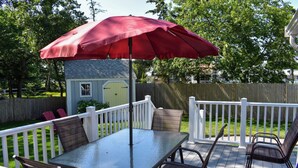 Een terras met een tafel en stoelen, een rode parasol en een klein tuinhuisje op de achtergrond.
