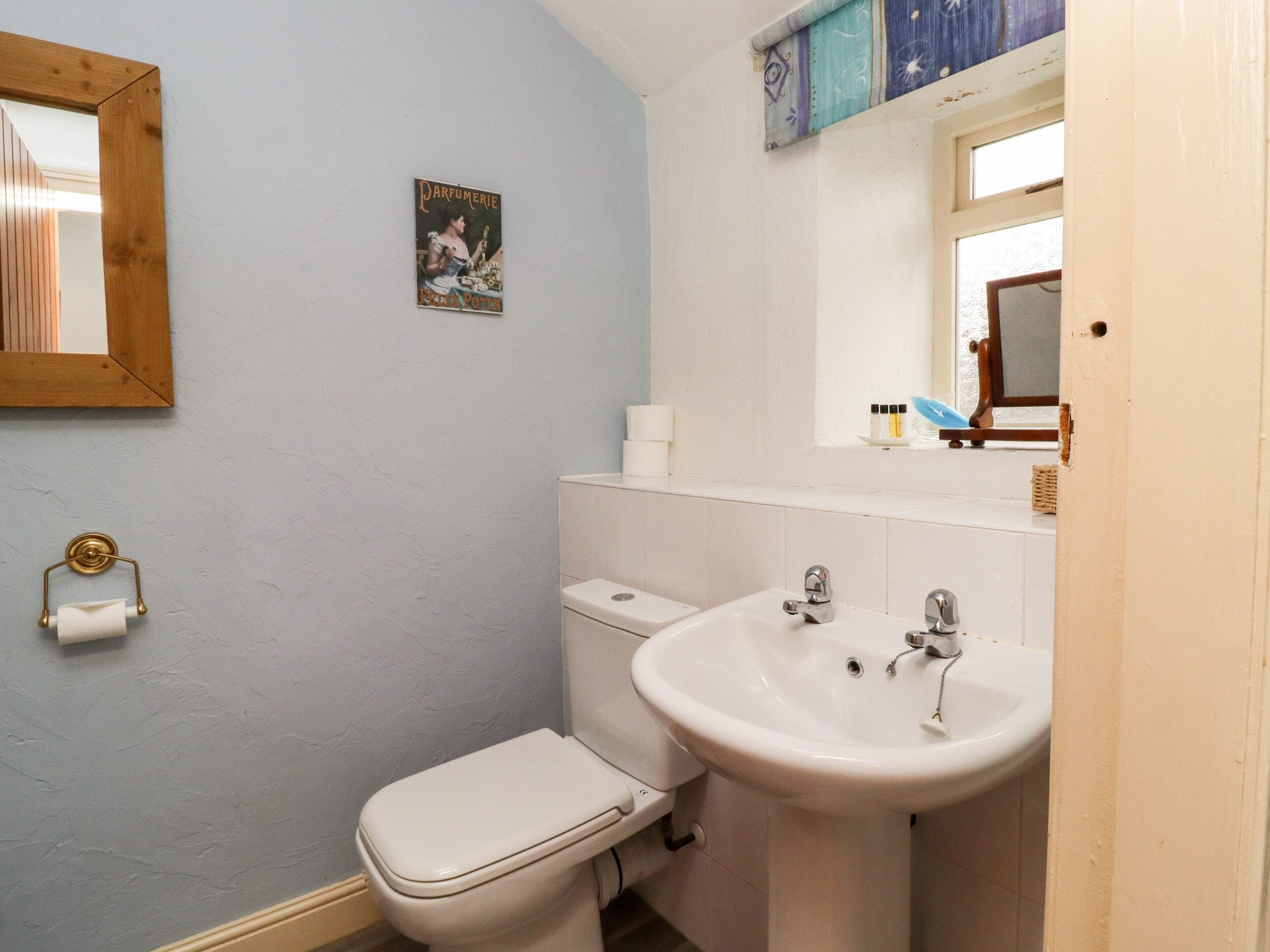 Un bagno con il water, un lavandino e uno specchio. C'è un poster appeso al muro e un rotolo di carta igienica.