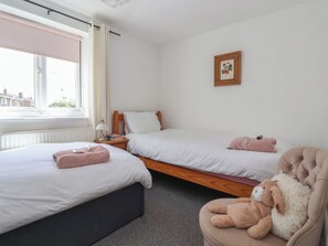 A bedroom with two beds, a window, a radiator, a bedside table with books, a teddy bear, and a framed botanical illustration.