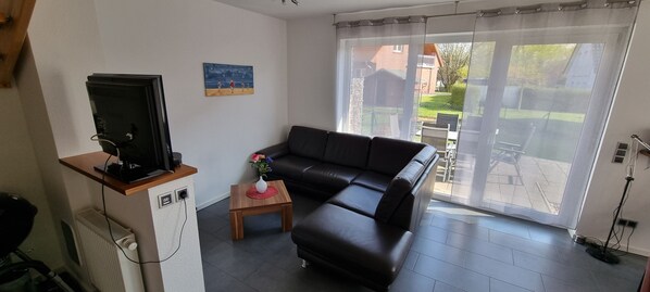 A living room with a black leather sofa, a small wooden table with a vase, a television on a wooden stand, and a sliding glass door leading to an outdoor area.