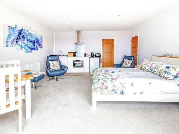 A modern bedroom with a bed, two chairs, a desk, a radiator, and a kitchen area. - Greenside Studio (Penzance)