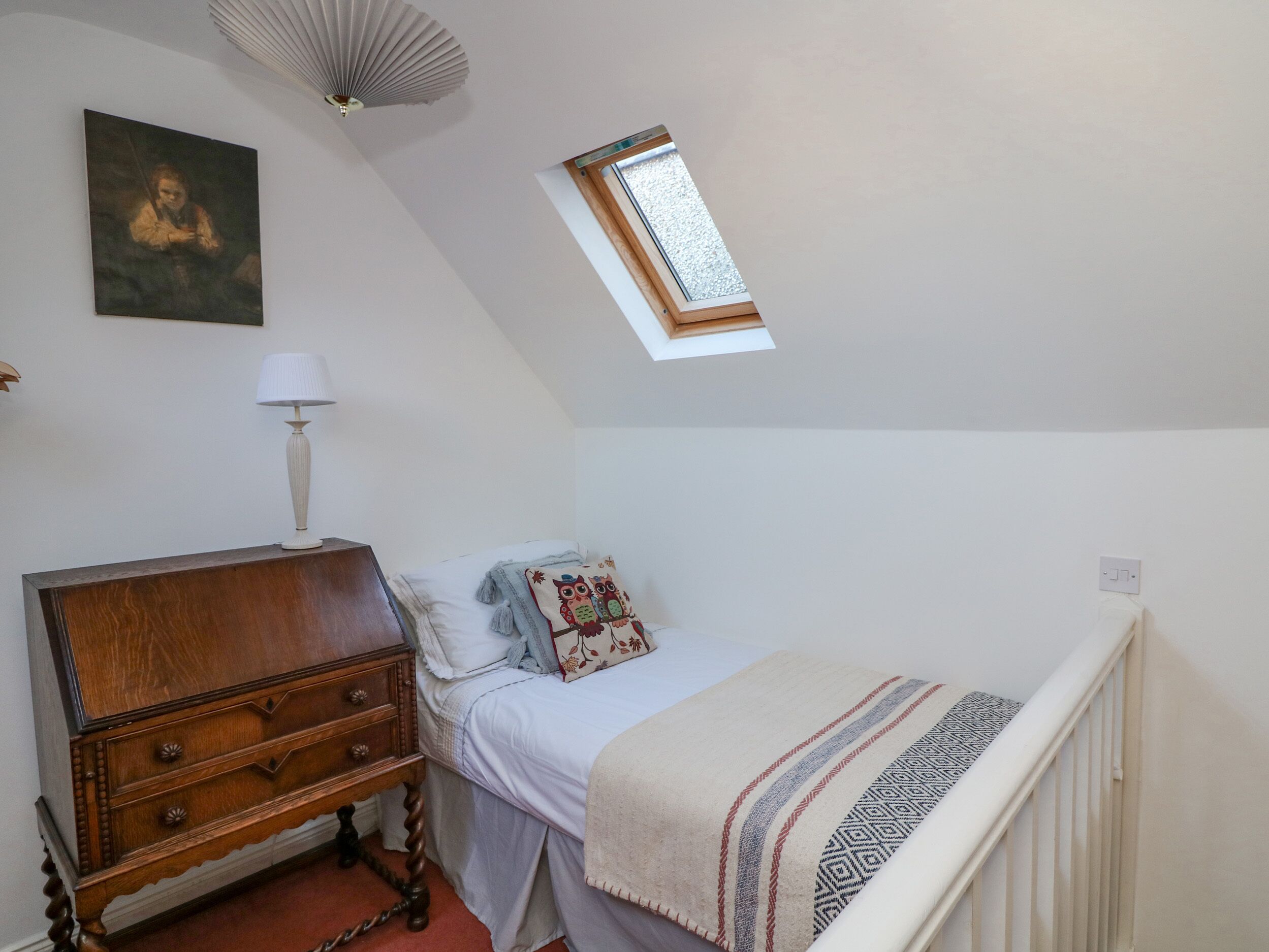A bedroom with a wooden dresser, a bed with a patterned pillow, a nightstand with a lamp, a ceiling fan, and a skylight.