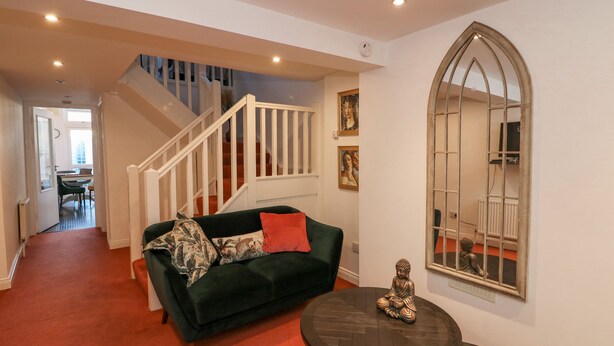 Ein Wohnzimmer mit einem dunkelgrünen Sofa, einem kleinen runden Tisch und einer Buddha-Statue. Eine Treppe mit weißen Geländern führt zu einem weiteren Raum.