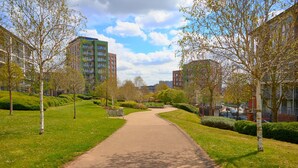 A pathway flanked by trees and buildings, leading to a park area with benches.
