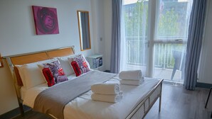 A neatly made bed with white linens and rolled towels, a bedside table with a lamp, a mirror on the wall, and a window with curtains.