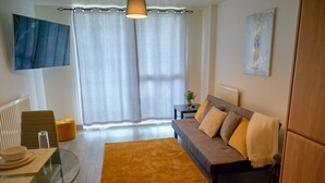 A modern living room with a glass dining table, a sofa with cushions, a yellow rug, and a large window with curtains.