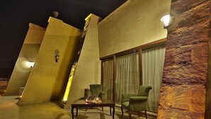 Un espacio exterior moderno con mobiliario, una mesa, sillas, un muro de piedra y un amplio ventanal con cortinas.