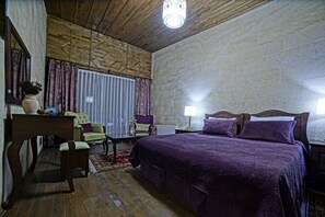 Une chambre avec un plafond en bois, un grand lit avec une literie violette, un fauteuil vert, un bureau en bois et une fenêtre avec des rideaux.
