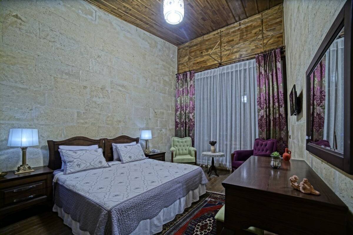 A bedroom with a large bed, two bedside tables with lamps, a green armchair, a purple armchair, a small round table with a vase, and a mirror on the wall.