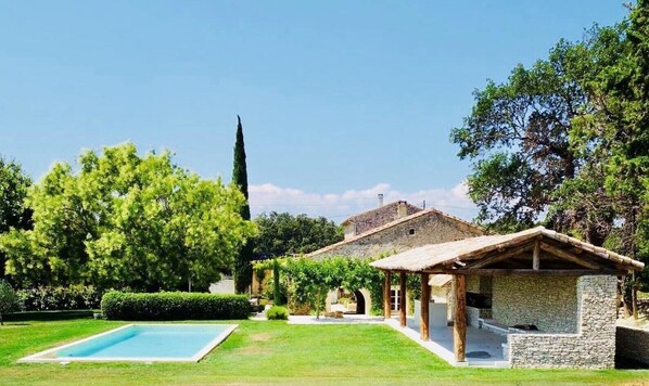 Une maison traditionnelle en pierre, dotée d’une piscine, entourée d’une végétation luxuriante et d’un grand arbre.