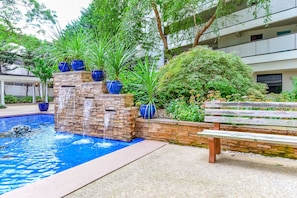 Una piscina con cascada, rodeada de plantas en macetas y un banco de madera.