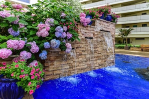Un hotel con pared de piedra, una fuente en cascada y una piscina con agua azul.