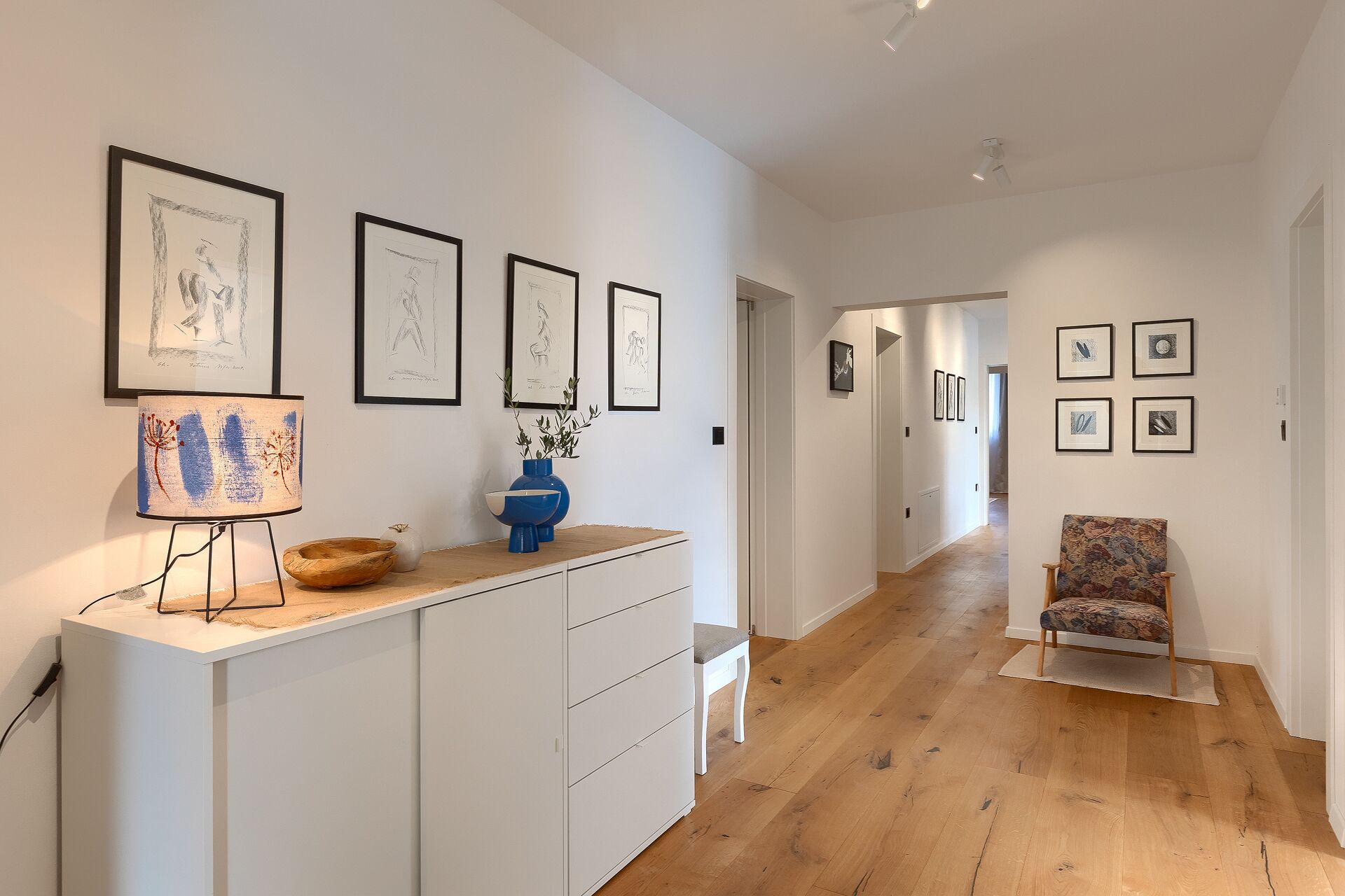 A hallway with framed artwork on the walls, a desk with a lamp and decorative items, and a chair in the corner.