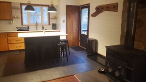 Een keuken met een centraal eiland, houten kastjes, een stenen open haard en een raam met uitzicht op een terras.