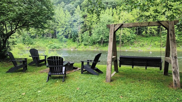 Een rustige buitenruimte met Adirondack-stoelen en een houten bank aan een rivier, omgeven door weelderige begroeiing.
