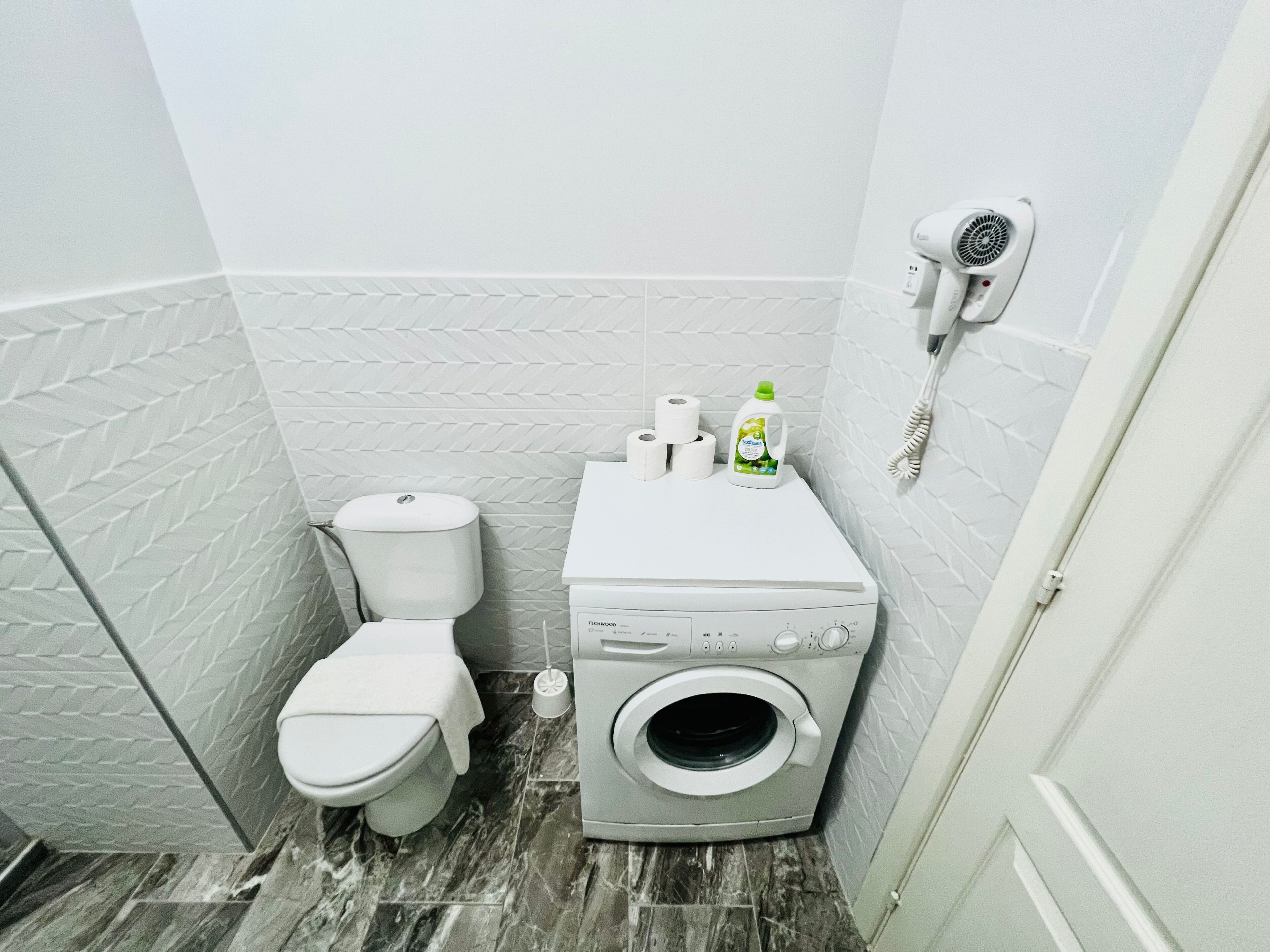 A compact bathroom with a toilet, washing machine, and a hairdryer on a wall-mounted rack.
