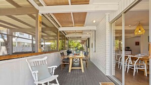 A covered outdoor patio with a wooden table and chairs, a rocking chair, and a glass door leading to an interior dining area. - Tulum Hideaway – Steps to Water, Close to Trails (Callala Bay)