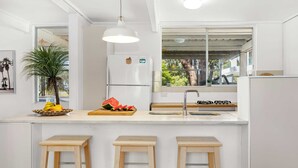 A modern kitchen with a central island, wooden stools, a refrigerator, and a window with a view of greenery. - Tulum Hideaway – Steps to Water, Close to Trails (Callala Bay)