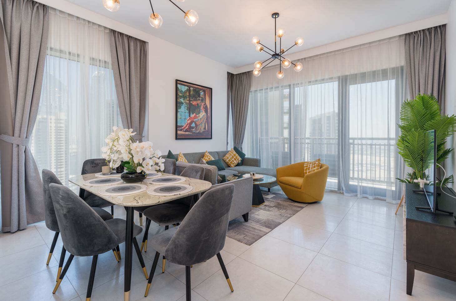 Een moderne woonkamer met een eethoek, grijze gestoffeerde meubels, een ronde tafel met een vaas witte bloemen en een groot raam met lichte gordijnen.
