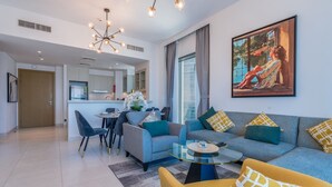Una sala de estar moderna con un sofá, sillones y una mesa de centro. Hay una zona de comedor con sillas y una mesa. La habitación tiene un ventanal con cortinas y un cuadro en la pared.