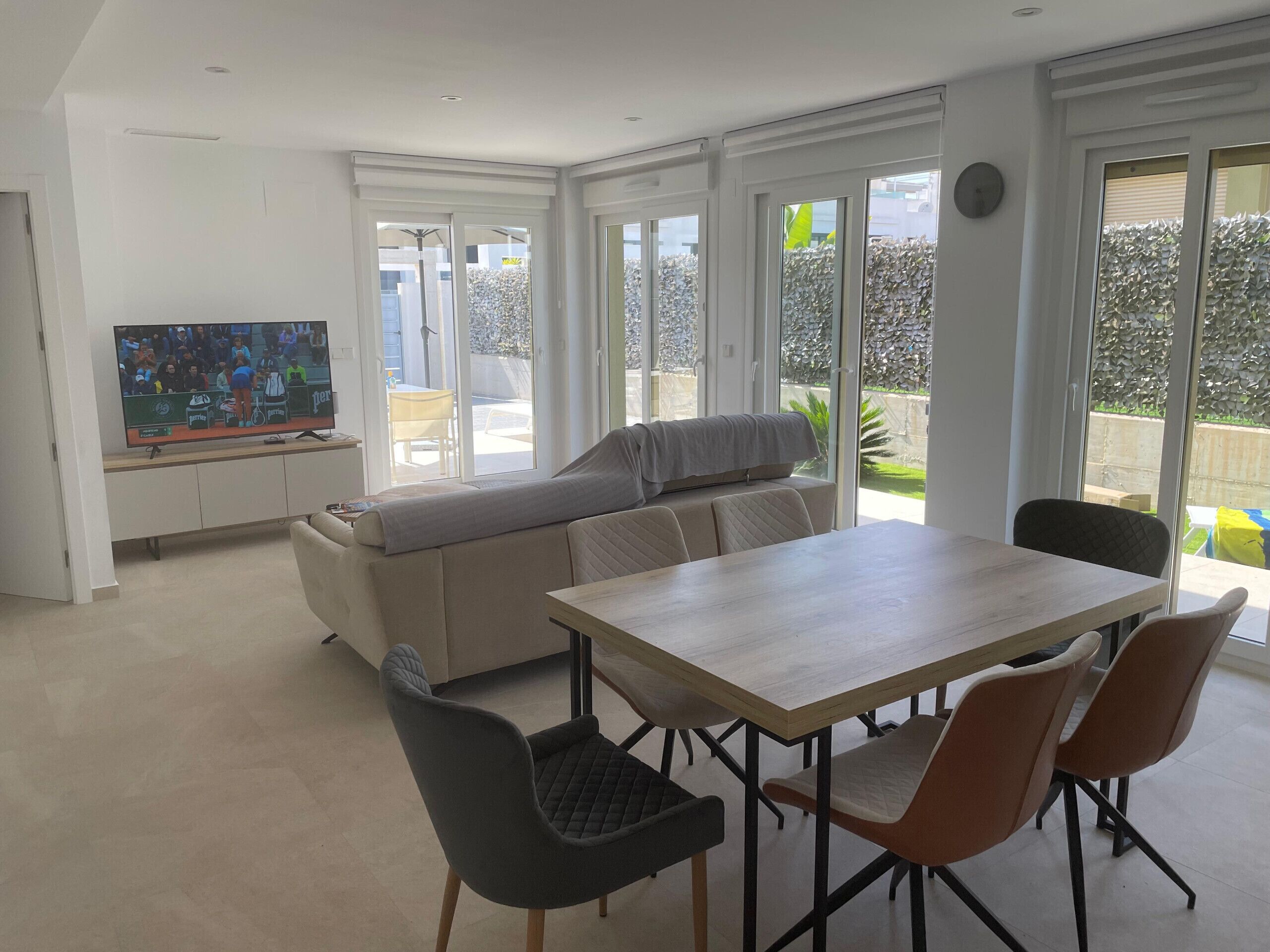 A modern living room with a dining area, large windows, and a television displaying a tennis match.