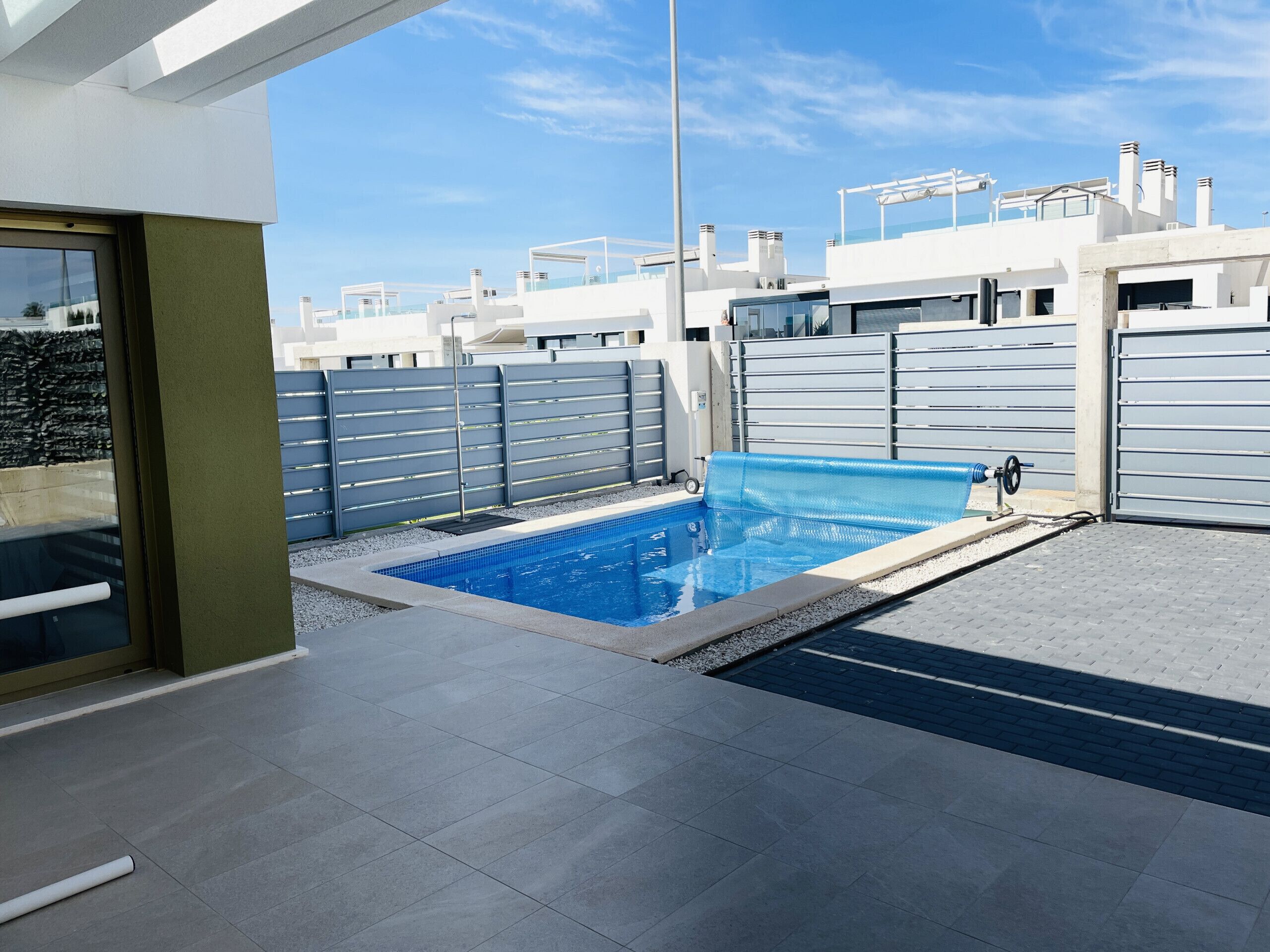 Une piscine extérieure moderne, dotée d’une structure en verre, entourée d’une clôture métallique et d’une terrasse dallée.