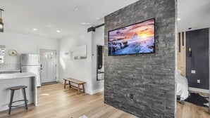 Moderni keittiö, jossa on valkoiset kaapit, puinen penkki ja suuri televisio, joka on kiinnitetty kiviseinään.
