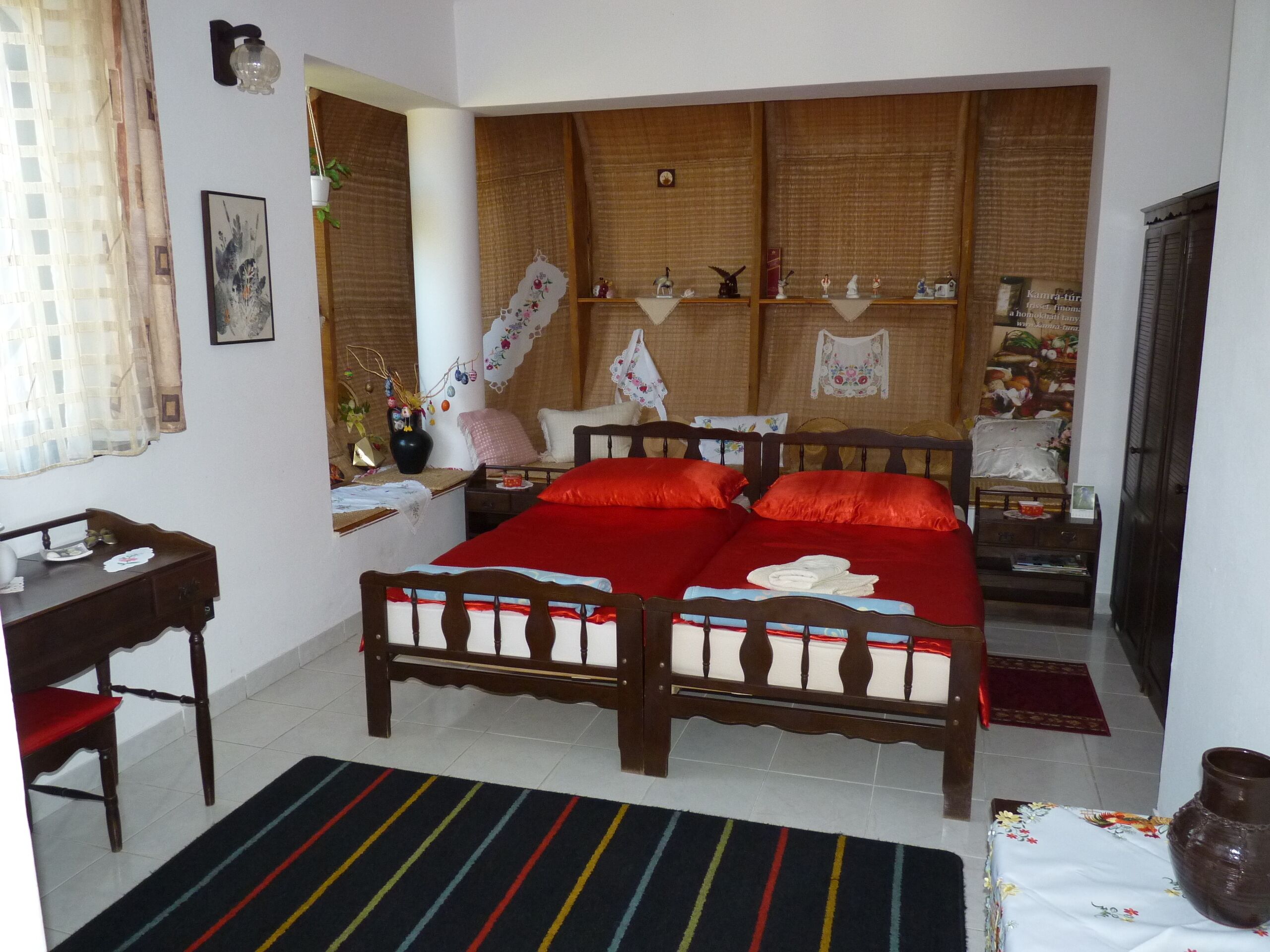 Un dormitorio con cama de madera, una mesita de noche, una silla, un ventanal con cortinas y un estante en la pared con objetos decorativos.