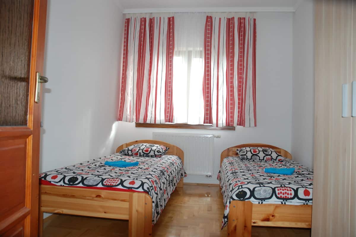 A small, well-lit bedroom with two wooden beds, a window with red curtains, and a radiator below.