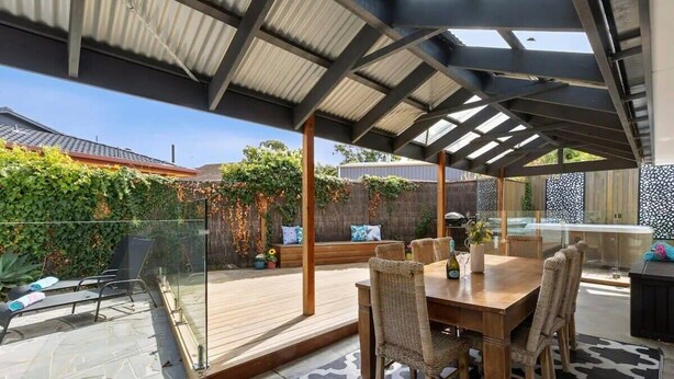 A covered patio with a wooden table and wicker chairs, a glass railing, and a patterned rug. - Sunny Daze Haven: SPA and Pool (Ocean Grove)
