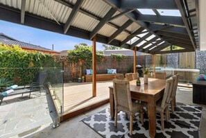 Een overdekt terras met een houten tafel en rieten stoelen, een glazen balustrade en een kamerbreed tapijt.