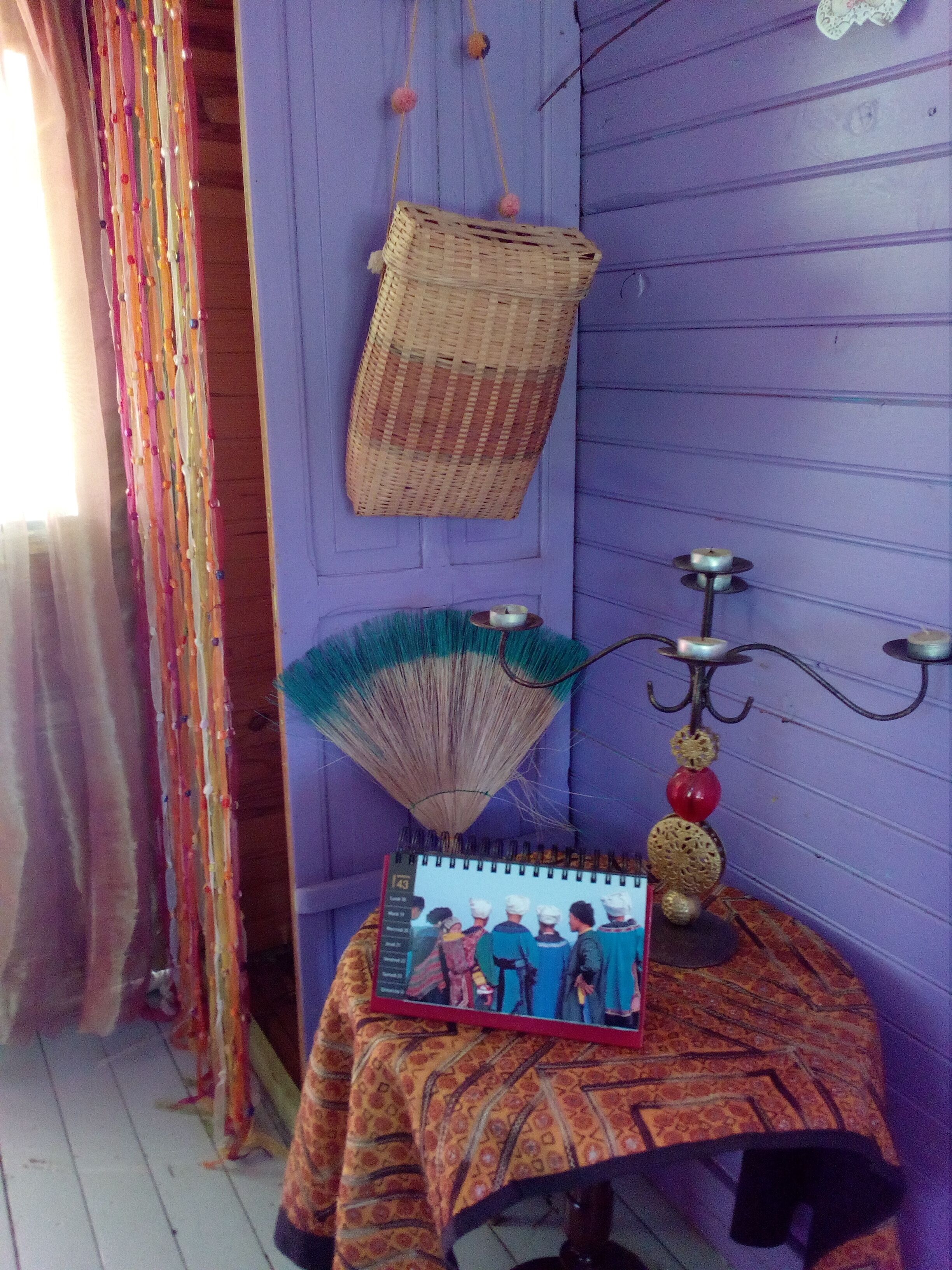 A room with a purple wall, a woven basket hanging, a fan, a candle holder, and a calendar on a table.