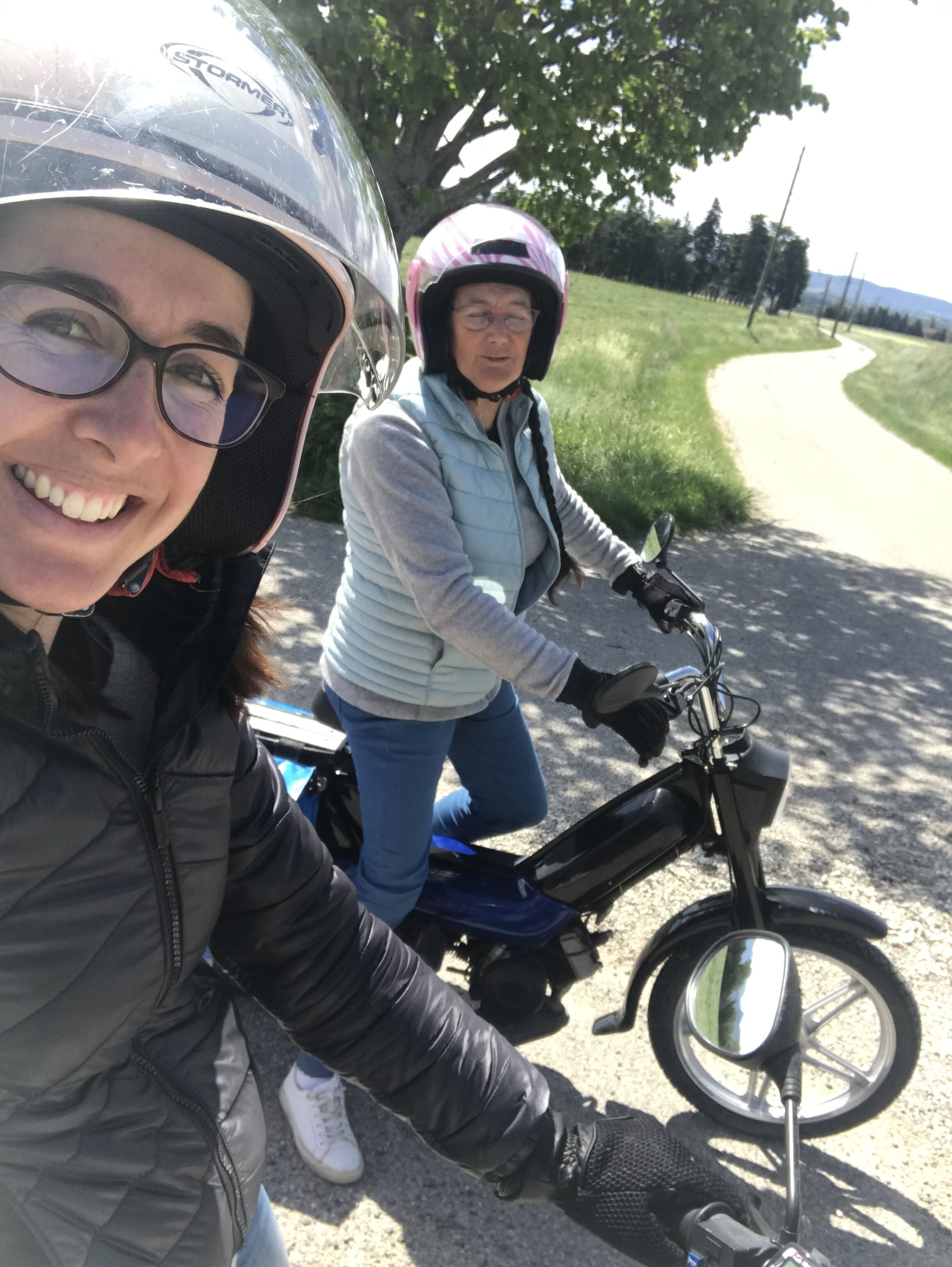 Zwei Personen fahren auf einem Roller, eine davon trägt einen Helm, im Hintergrund ein asphaltierter Weg.