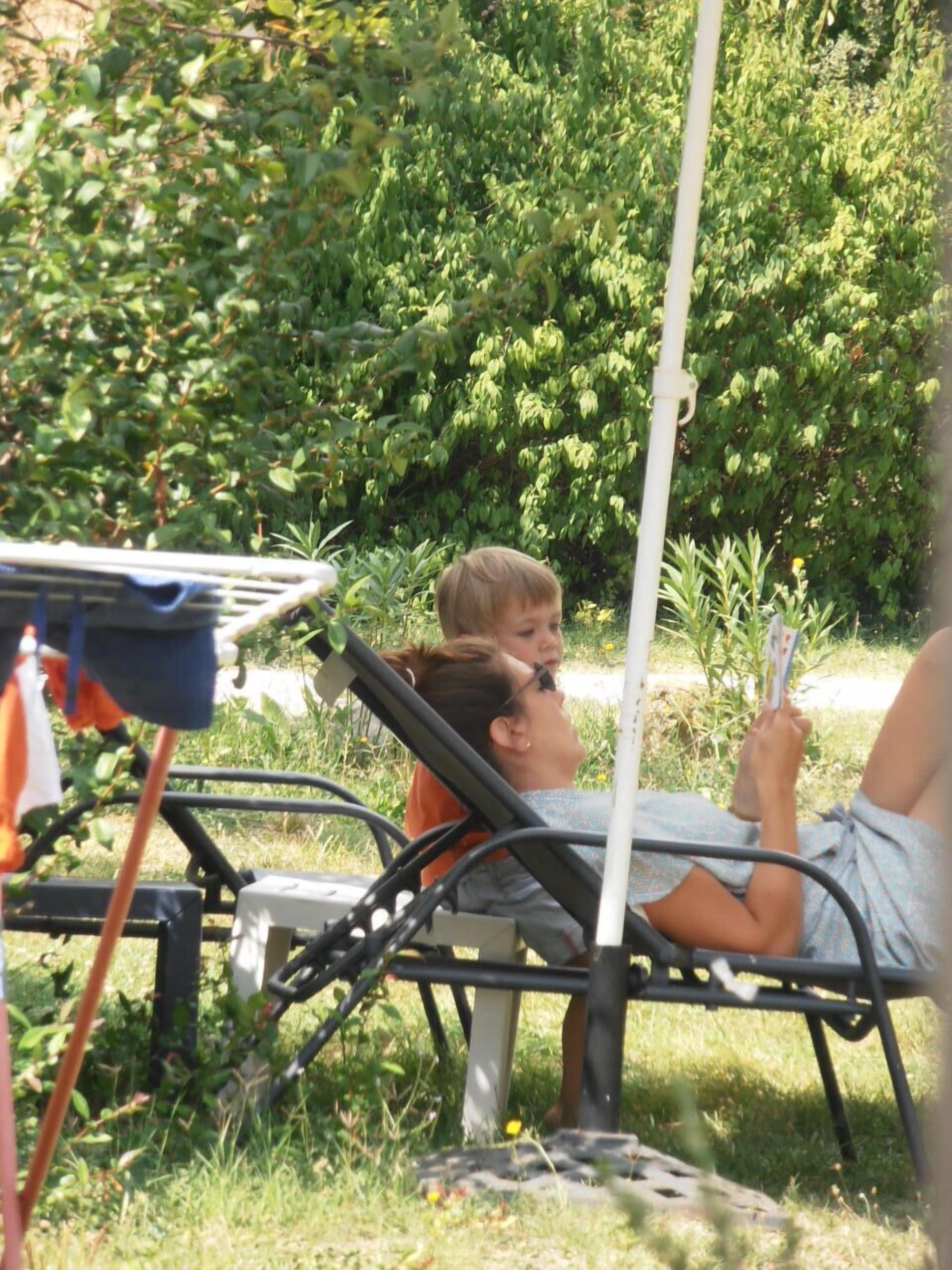 A person lying on a metal chair outdoors, surrounded by greenery and a white pole.