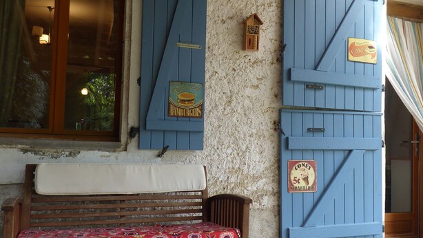 Un banco de madera con un cojín floral, una mesa con una jofaina y vasos, y contra una pared texturizada, contraventanas azules.