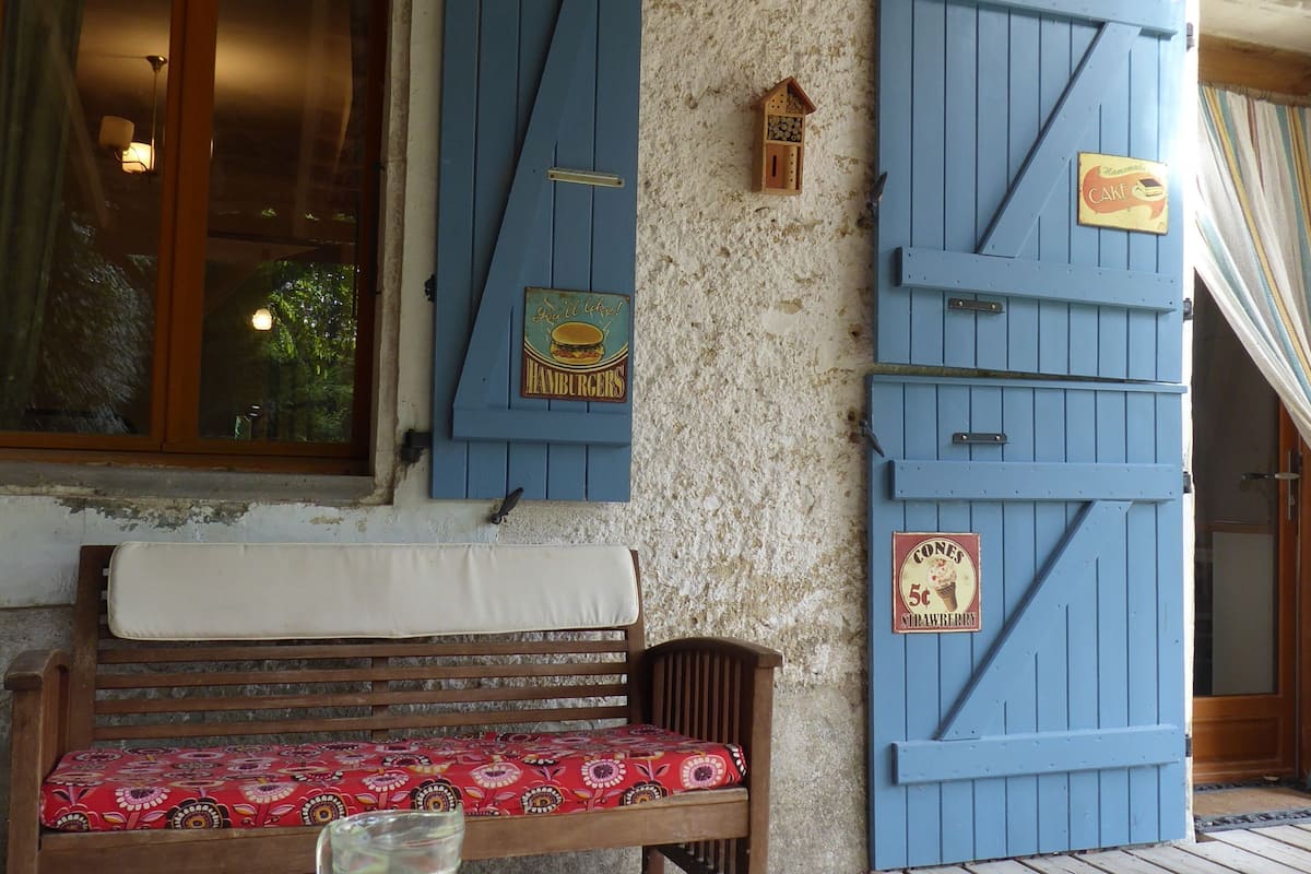 A wooden bench with a floral cushion, a table with a pitcher and glasses, and blue shutters on a textured wall.