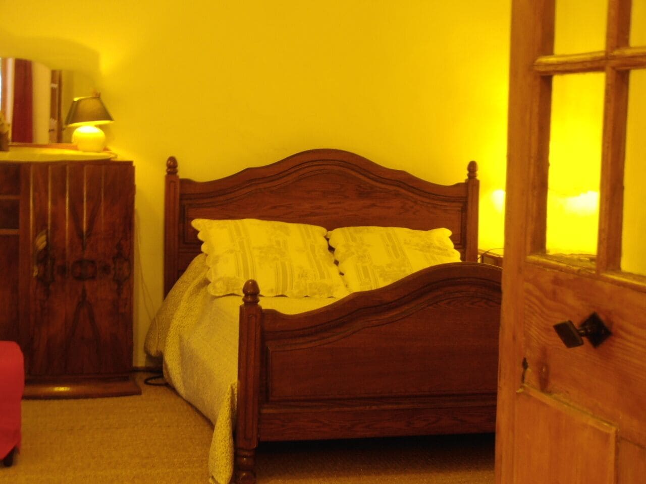 Ein Bett mit Holzkopfstück, ein Nachttisch mit Lampe und ein Kleiderschrank in einem Zimmer mit gelben Wänden.