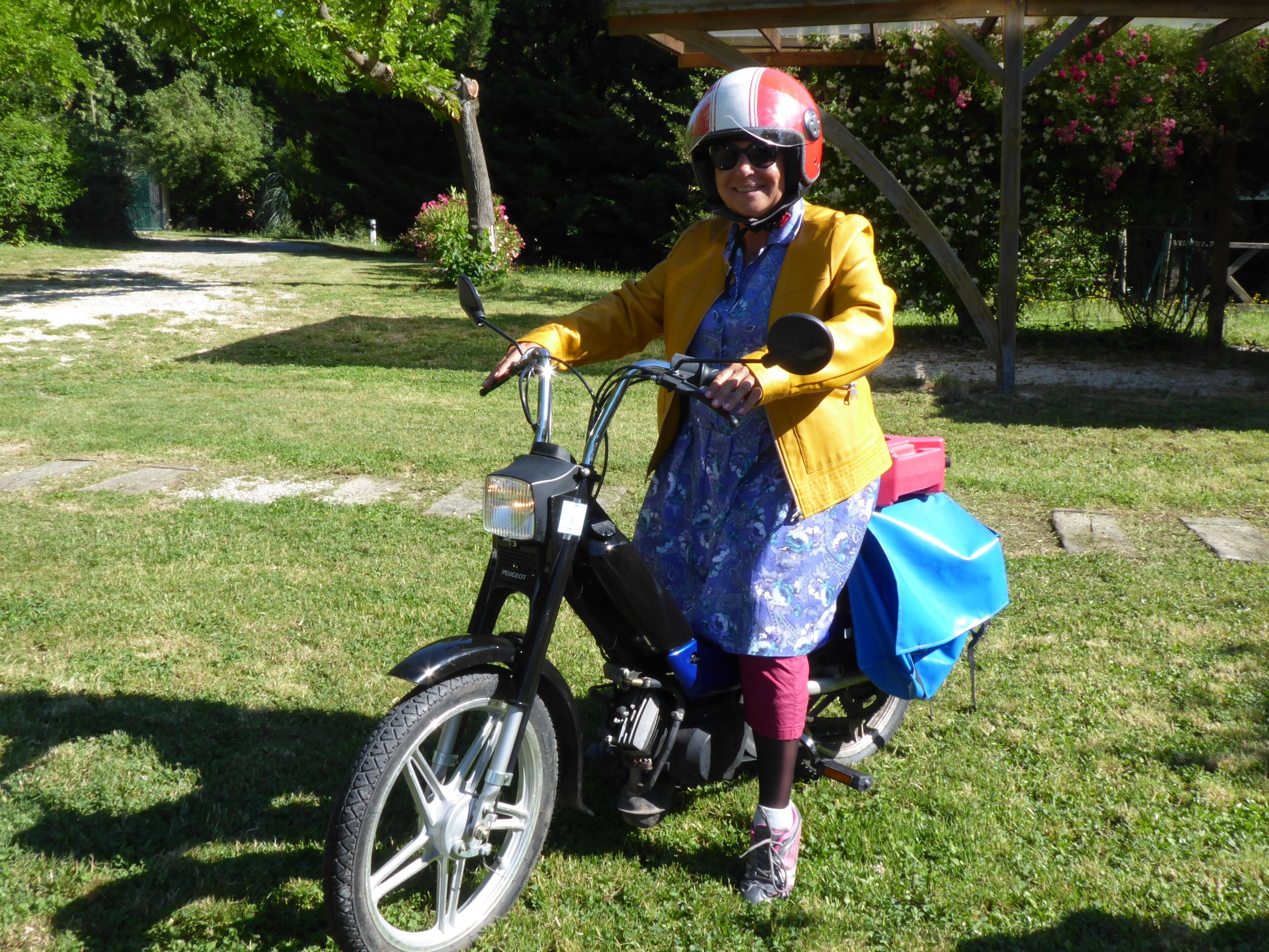 A person on a black bicycle with a blue bag, wearing a yellow jacket and a helmet, in a grassy outdoor area with trees and a wooden structure.