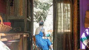 Una habitación rústica de madera con una cama, una mesa y una silla. Hay una ventana con cortinas y vistas a árboles afuera.