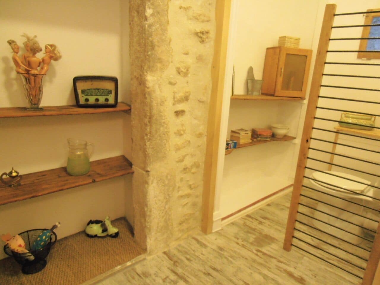 Ein Zimmer mit rustikaler Steinwand, Holzregalen mit diversen Gegenständen, einem kleinen Tisch mit einem Krug und einem Holzkäfig mit einem WC.