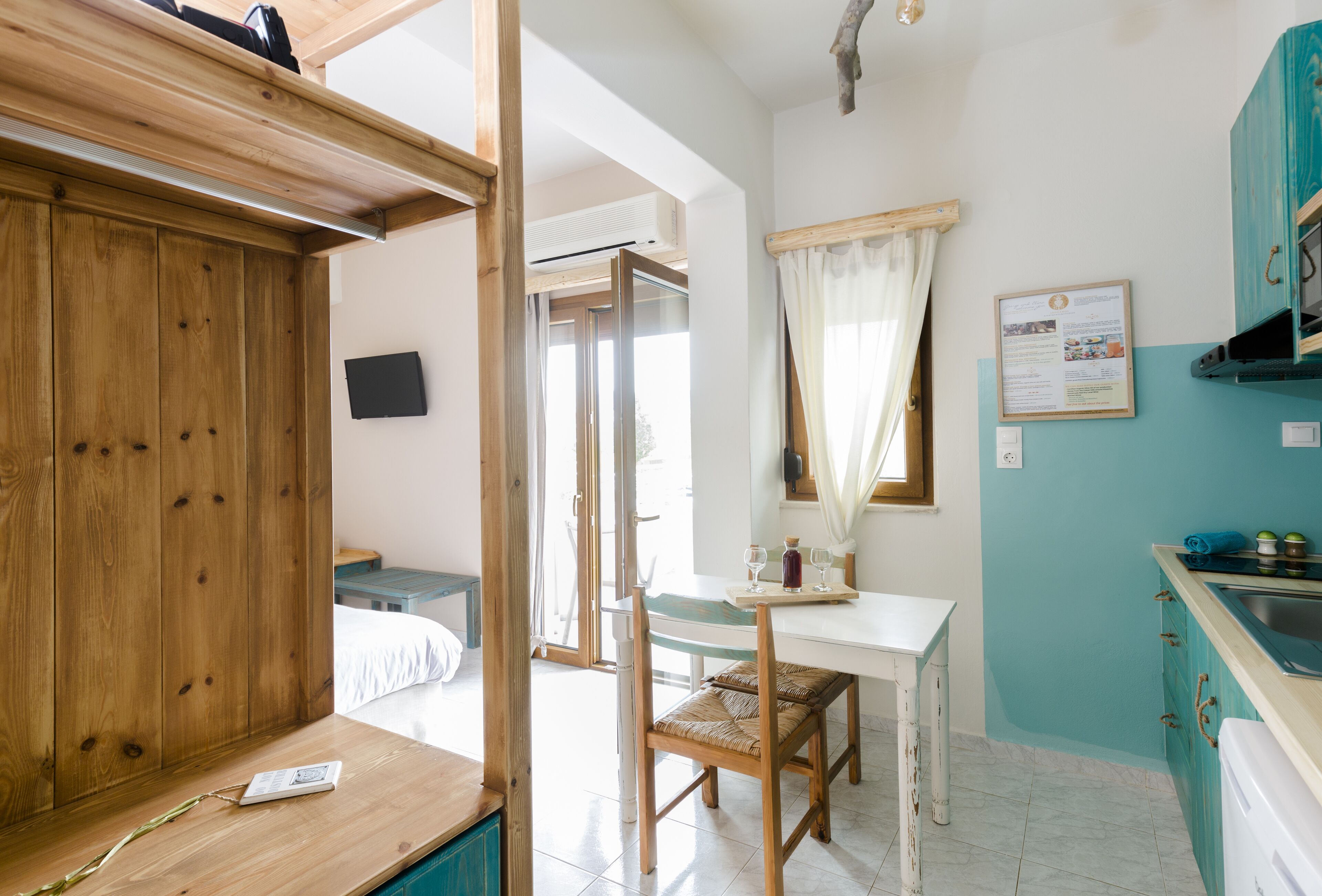 A room with a wooden wardrobe, a dining table with chairs, a kitchen area with a sink, and a window with curtains.