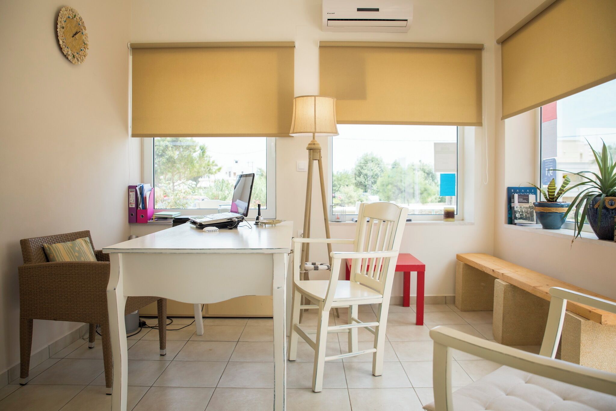 A room with a desk, laptop, two chairs, a lamp, a bench, and a window with blinds.
