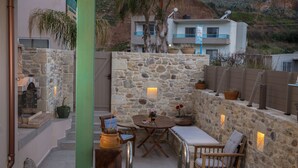 Un espacio al aire libre con paredes de piedra, una mesa y sillas, rodeado de plantas en macetas y un pilar verde.