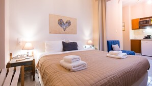 Una habitación de hotel con una cama grande, una mesita de noche con lámpara, una silla y una cocina visible a través de una puerta abierta.