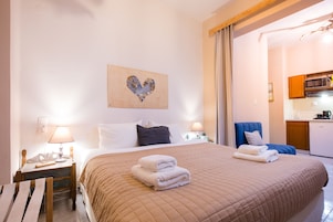 Una habitación de hotel con una cama grande, una mesita de noche con lámpara, una silla y una cocina visible a través de una puerta abierta.
