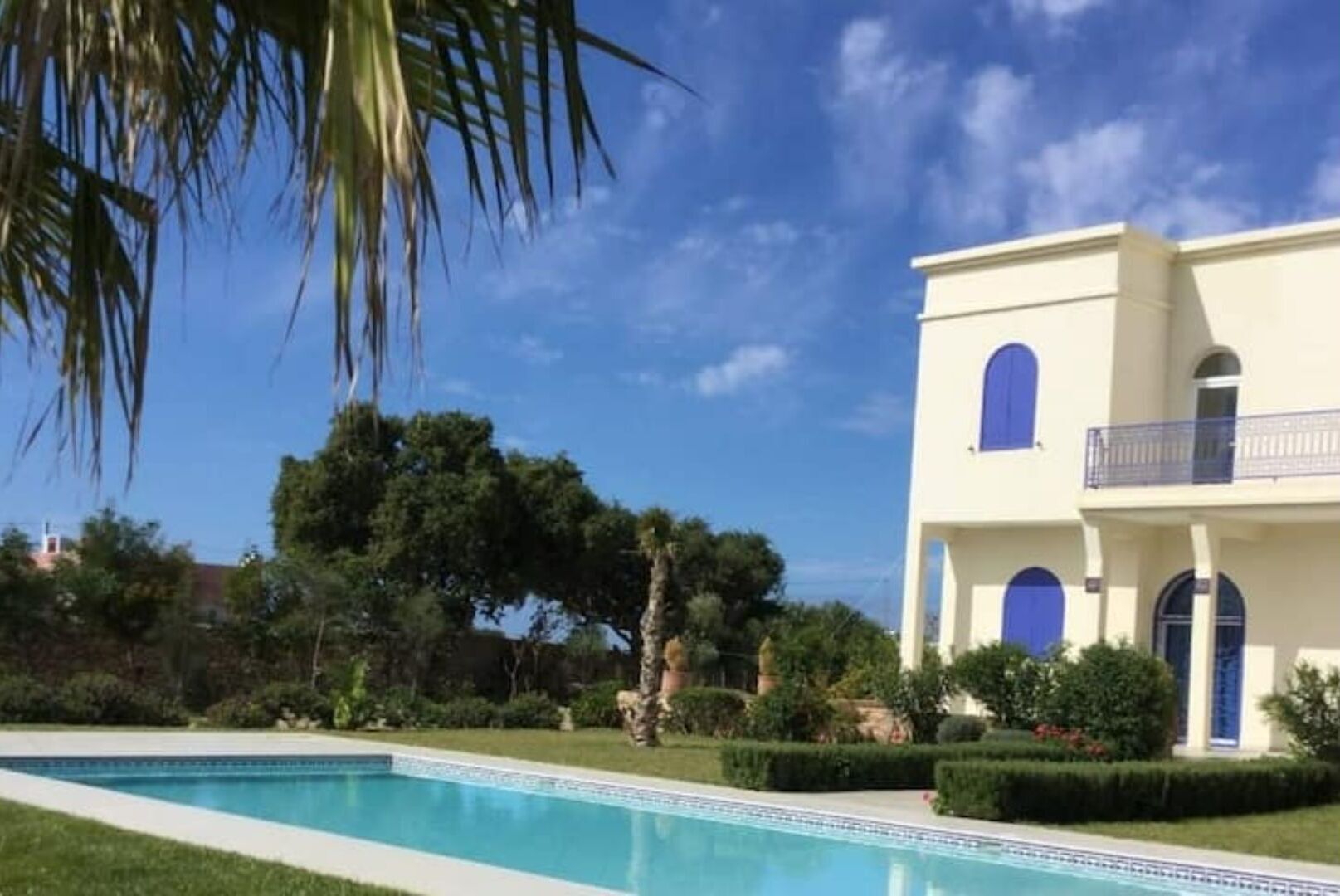 Blick auf einen Pool vor einem zweistöckigen Gebäude mit Balkon, umgeben von Grün und blauem Himmel.